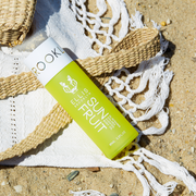 Sun Fruit Golden Glistening Scented Body Oil 95ml resting on white tasseled towel and beige straw bag on sand.