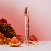 West 10ml Eau De Parfum Travel Spray with cap removed on white table next to blood orange wedges and orange sunflower on pink gradient background.