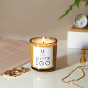Superego 6.5 oz. Terrific Scented Candle with wick lit on white table near open book, gold earrings gold necklace, wooden chess piece, in front of white box with wooden top with compass and green plant resting on top, against on beige background.