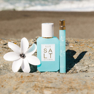 Salt 50ml Eau De Parfum between Salt 10ml Eau De Parfum Travel Spray and white flower on rock with a beach in the background.